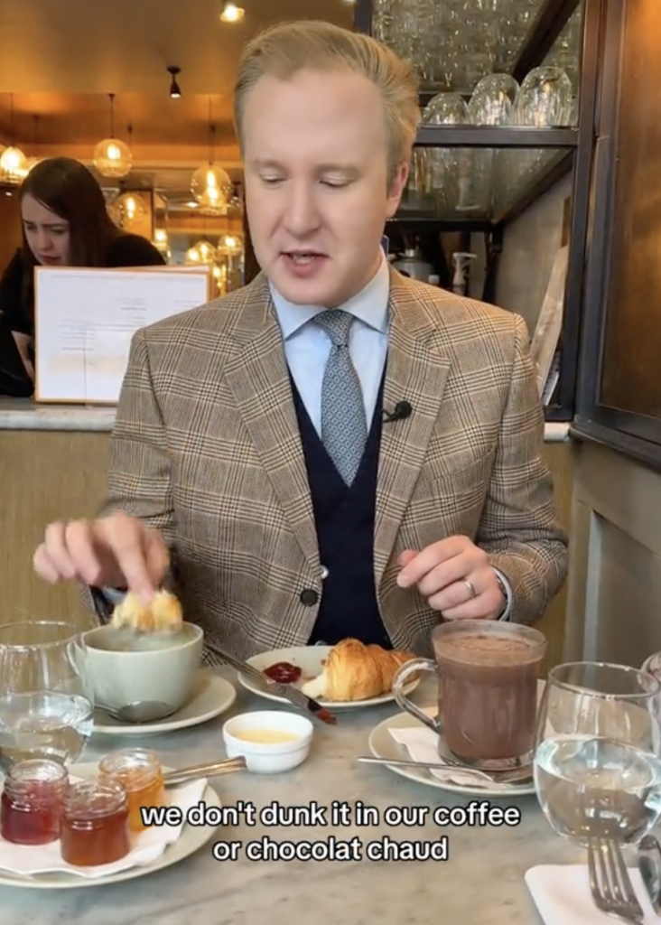 British etiquette expert William Hanson sitting at a table with a croissant, coffee, hot chocolate etc.
